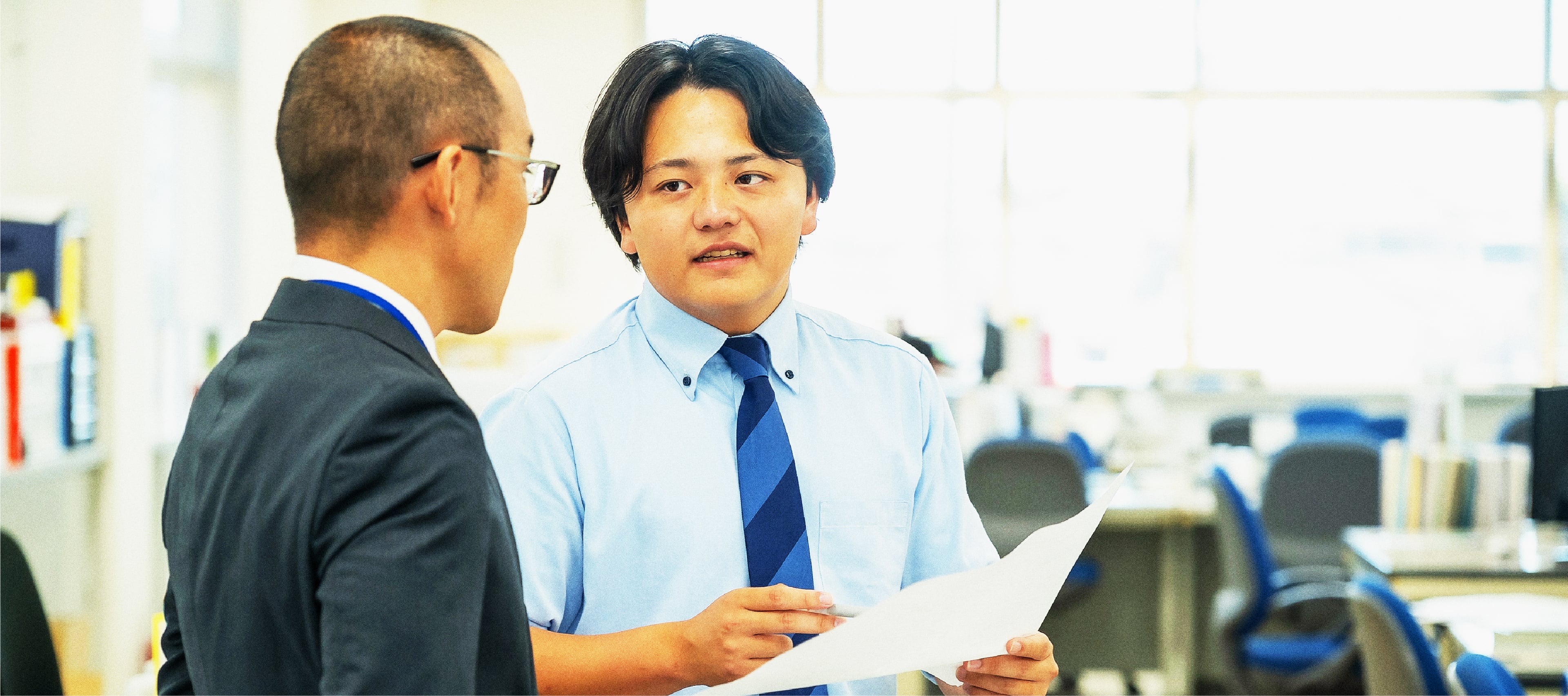オフィス内で会話するスーツ姿のビジネスマンとネクタイを締めた男性